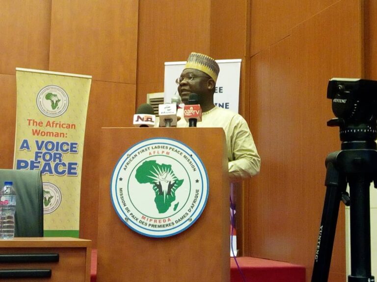 A man wearing white khaftan on a podium holding a microphone, with a banner in his background with peace inscribed on it